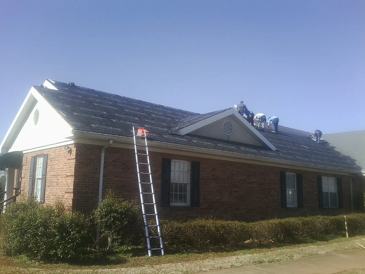 Crew tearing shingles off a brick ranch roof with a ladder set — Metal Roof Repair work by our crew in Center Conway