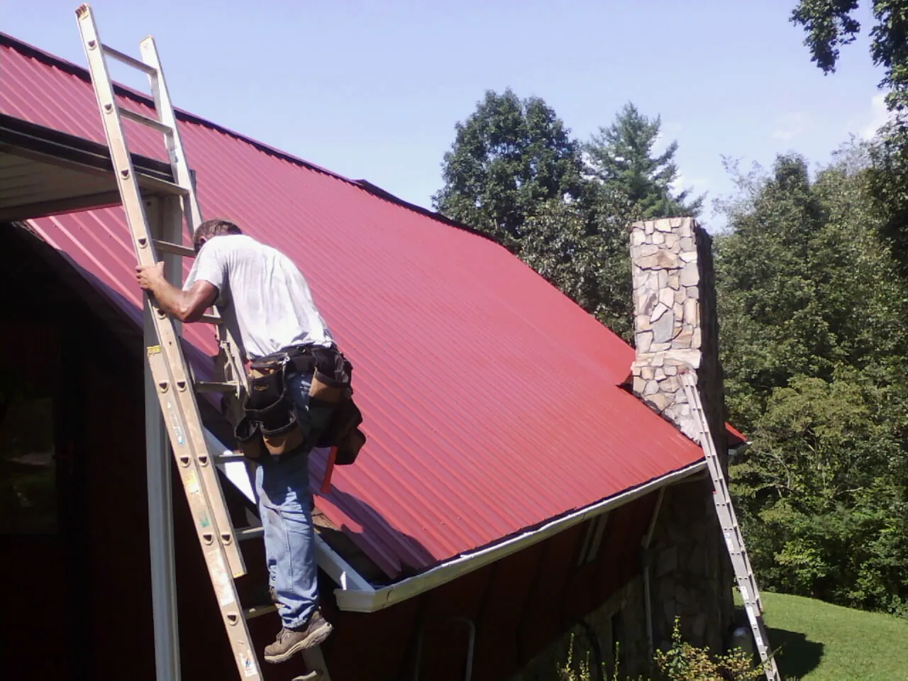 Roofer on a ladder beside a red metal roof and stone chimney — Soffit & Fascia Repair by Roofing Brothers in Center Conway